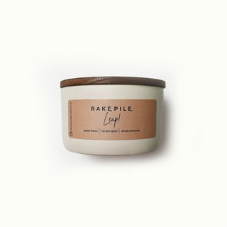 Candle with a wooden lid and 'RAKE, PILE, LEAP!' label on a white background