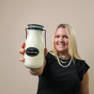 Person holding a 14 oz glass milk bottle with a 'Oatmeal, Milk & Honey' label against a plain background