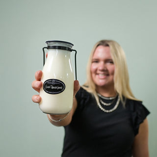 Person holding a glass 14 oz milk bottle candle with a 'Sweet Tobacco Leaves' label, against a plain background
