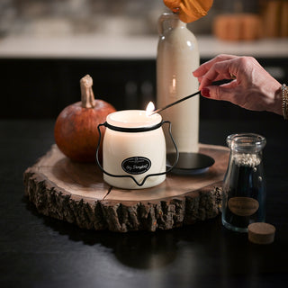 Person lighting a 16 oz 'Hey, Pumpkin!' candle in a white glass container with pumpkins and vase in the background