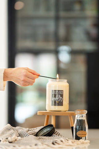 A 22oz 'Wildberry Waffles' candle being lit on a wooden stand with a bottle of matches on a white surface with a blanket
