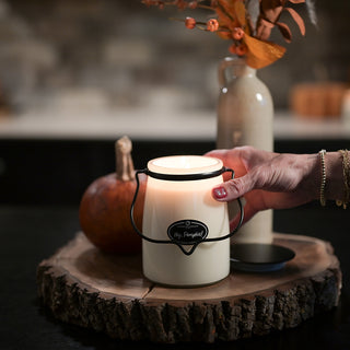 Hand holding a lit 22 oz 'Hey, Pumpkin!' candle on a wooden surface with pumpkins and a vase in the background