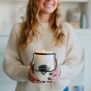 Person holding a lit 22 oz candle with a 'Oatmeal, Milk & Honey' label and black metal handle