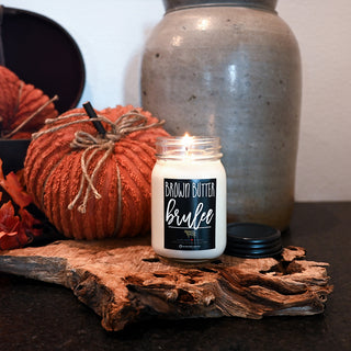 Candle labeled 'Brown Butter Brulee' on a wooden surface with pumpkins and a jar in the background.