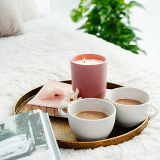 Lit candle in a blush colored ceramic container sitting on a book with crystals and flower in a tray with cups of coffee on a blurred white surface