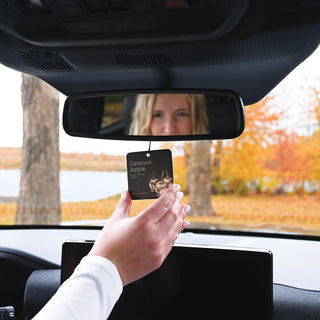 Person holding a car air freshener in front of a car's rearview mirror with autumn scenery outside.