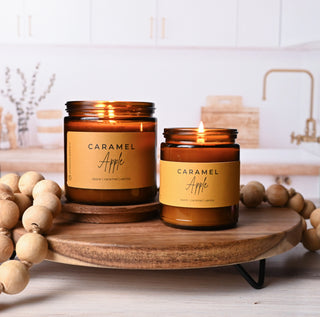Two amber candle jars in different sizes lit, sitting on a wooden riser with wooden beads in a kitchen setting.