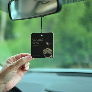 A hand holds a square car air freshener tag labeled 'Caramel Corn' in front of a car windshield, with a rearview mirror and blurred green outdoor scenery in the background.