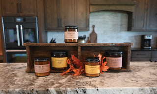 Four glass candle jars with black lids and pastel labels arranged on a rustic wooden crate on a kitchen countertop. The setting includes a kitchen background with wooden cabinets and a stove, and decorative orange autumn leaves are placed in front of the jars.
