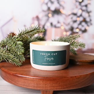 Candle labeled 'Fresh Cut Fraser' on a wooden surface with Christmas tree branches.