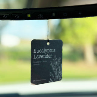 A rectangular car air freshener tag labeled 'Eucalyptus Lavender' hanging from a windshield wiper arm inside a vehicle, with a blurred outdoor background.