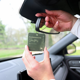 Person placing a car air freshener labeled 'Farmhouse Christmas' on a car's rearview mirror.