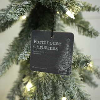 A black hanging tag for a 'Farmhouse Christmas' artificial tree, featuring white text and a snow-covered tree graphic, shown hanging from a flocked green Christmas tree with warm white string lights.