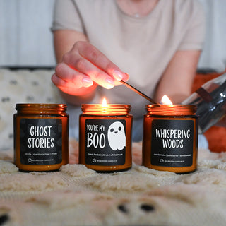 Three lit amber glass jar candles with black labels are arranged on a cream knit blanket. A person's hand holds a match near the flames.