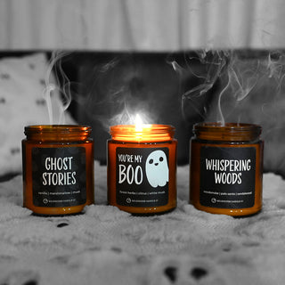Three Halloween-themed candles with labels 'Ghost Stories', 'You're My Boo', and 'Whispering Woods' on a textured surface.