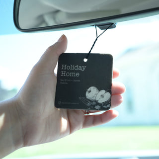 A hand holding a black car air freshener tag labeled 'Holiday Home' under a car rearview mirror.
