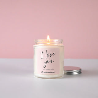 Lit candle in a clear jar with a silver lid sitting next to it, labeled 'I love you.' on a light pink label against a light pink background