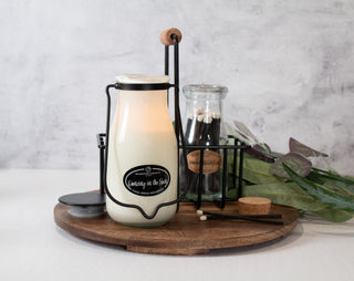 Lit candle in a milk-bottle sitting on a wooden tray with a black metal holder and match bottle against a marbled background