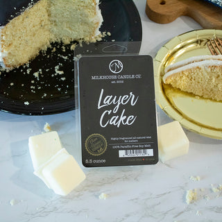 A packaged wax melt labeled 'Layer Cake' by Milkhouse Candle Co. sits on a marble surface with cake pieces and wax chunks around it.