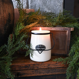 Candle in a white container with a black label on a wooden surface surrounded by greenery