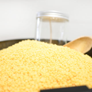 Yellow wax beads in a bowl with a wooden spoon and blurred jar in the background