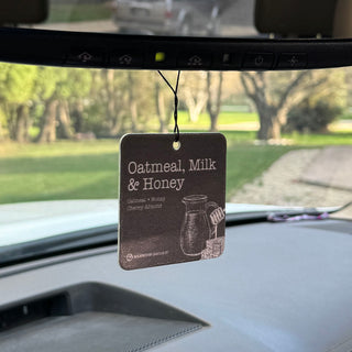 A car air freshener hanging from the rearview mirror inside a vehicle. The rectangular tag reads 'Oatmeal, Milk & Honey' and shows a simple brown illustration of a pitcher and cup, with a blurred outdoor scene visible through the windshield.