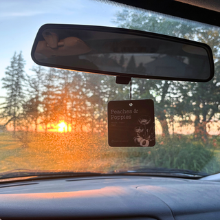 A car air freshener hanging from the rearview mirror, with the text 'Peaches & Poppies' visible on the tag, photographed from inside a car with a sunset scene outside.