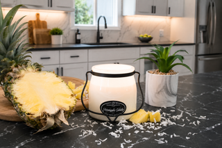 A white jar candle with a black metal handle and a pineapple-themed label reading 'Pineapple Gelato' sits on a dark marble kitchen countertop, surrounded by pineapple slices, a whole pineapple, and a small potted plant in a modern kitchen setting.