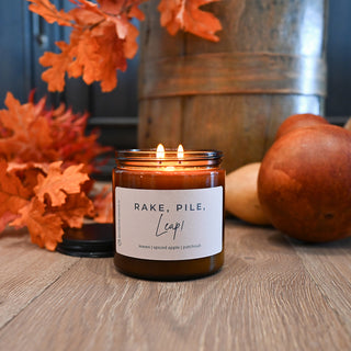 Candle in a jar labeled 'Rake, Pile, Leap' with autumn decorations on a wooden surface.