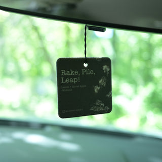 A square car air freshener hanging from a windshield wiper inside a vehicle, with a blurred green outdoor background. The tag reads 'Rake, Pile, Leap!' and 'Lavender + Sage + Apple + Peppermint' with botanical illustrations.