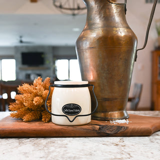 Small white container with black lid on a wooden cutting board next to a large bronze vase.