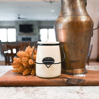 Small white container with black handle on a wooden cutting board next to a large bronze vase.