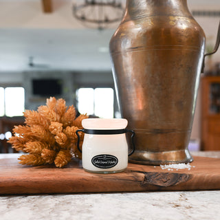 Small white container with black lid on a wooden surface next to a large bronze vase and dried plants.