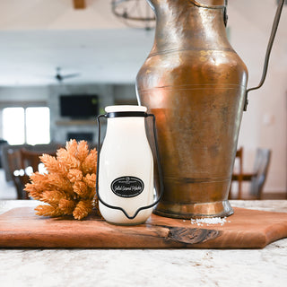 White container with black lid on a wooden cutting board next to a large bronze pitcher.