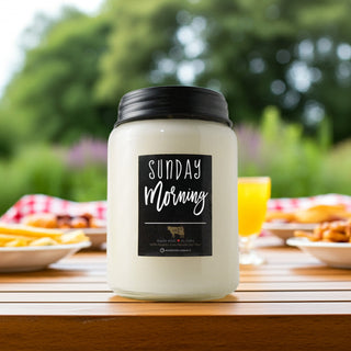 Candle labeled 'Sunday Morning' on a table with breakfast items in the background