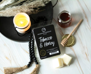 A pack of wax melts labeled 'Tobacco & Honey' by Milkhouse Candle Co. is styled in a flat lay with a lit candle, honey jar, wooden dipper, and a black beaded strand on a white marble surface.