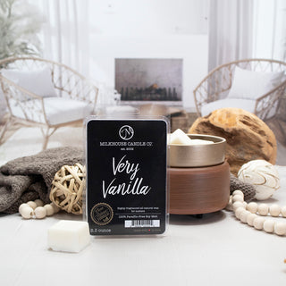 A packaged wax melt product labeled 'MILKHOUSE CANDLE CO. Very Vanilla' stands in the center of a styled interior scene with a lit candle, wooden bowl, beads, folded blanket, and neutral decor in the background.
