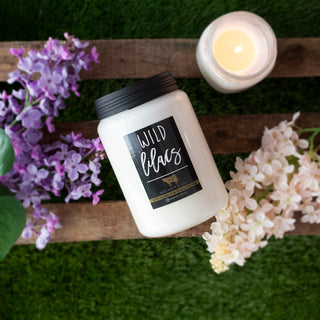 Candle labeled 'Wild Lilacs' on a wooden surface with flowers and a lit candle in the background