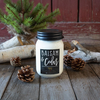 Candle labeled 'Balsam & Cedar' on a wooden surface with pinecones and a small tree