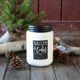 Candle labeled 'Balsam & Cedar' on a wooden surface with pinecones and a small tree