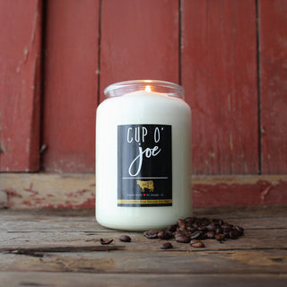 Candle labeled 'Cup O' Joe' with coffee beans on a wooden surface and red wooden panel background