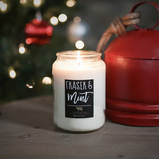 Candle with 'Fraser & Mint' label in front of a Christmas tree with lights and ornaments.