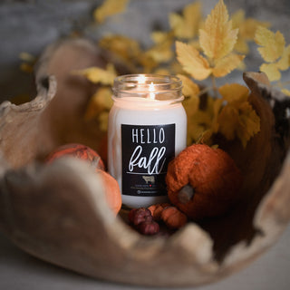 Candle labeled 'Hello Fall' in a decorative setting with pumpkins and leaves