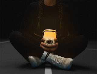A person sitting holding a lit 22 oz 'Linen & Ashwood' candle against a black background 