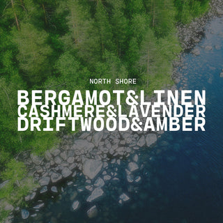 Scenic view of a forested area with a body of water, scent name of North Shore, with a text overlay of bergamot & linen, cashmere & lavender and driftwood & amber