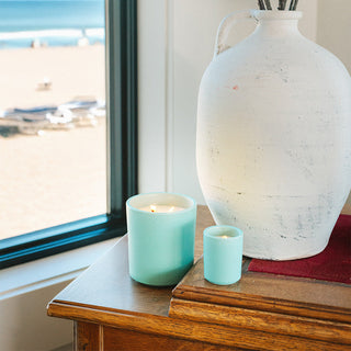 Two seafoam green candle container with lit candles sitting on a wooden table with a vase and blurred beach background