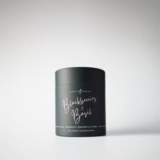 A black, cylindrical package containing a 'Blackberries & Basil' candle, with light pink writing, indicating it is a soy wax candle from the Earth & Ember line