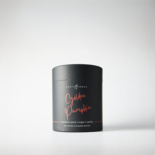 A black, cylindrical package containing a 'Golden Pumpkin' candle, with orange writing, indicating it is a soy wax candle from the Earth & Ember line