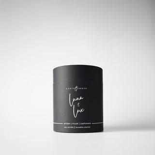 A black, cylindrical package containing a 'Luna & Lux' candle, with white writing, indicating it is a soy wax candle from the Earth & Ember line