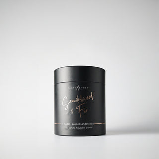 A black, cylindrical package containing a 'Sandalwood & Fir' candle, with rose gold writing, indicating it is a soy wax candle from the Earth & Ember line
